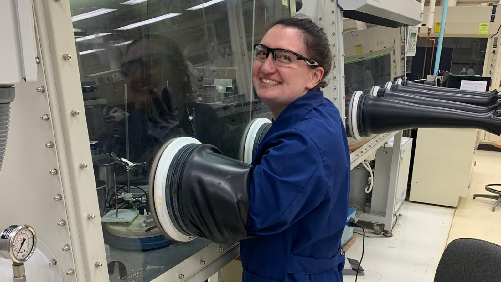 Becca Segel in a lab wearing goggles and a blue lab coat smiling at the camera